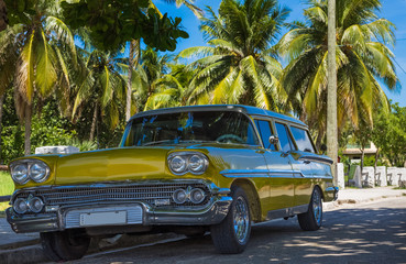 Amerikanischer gold gelber Oldtimer parkt in Varadero nahe des Strandes unter Palmen in Kuba - Serie Kuba Reportage