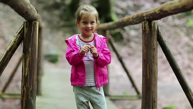 Funny little girl in pink jacket on the nature. Little girl stands on a wooden bridge and does dance moves