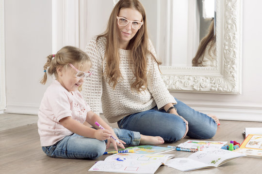 The daughter and mother sit on the floor and draw. The woman helps to the child to draw with markers. 