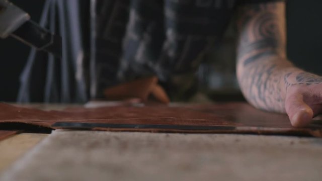 Craftsman's hands cutting a piece of leather on the table with a knife, dolly shot, close up