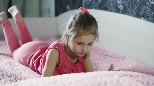 Little girl lying on the bed and playing computer games on the tablet.