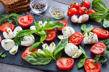 Cherry tomatoes, mozzarella cheese, basil, pesto sauce, dark rye grain bread and spices on black slate stone chalkboard. Italian traditional caprese salad ingredients. Mediterranean food. Top view
