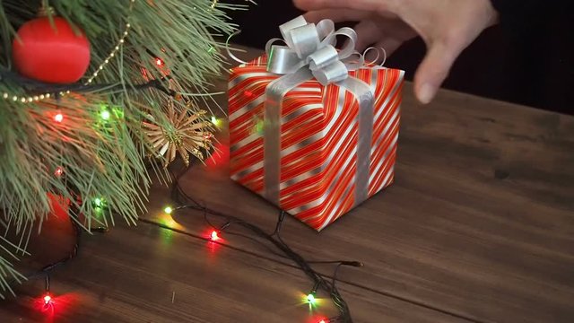 The man puts gift boxes under the Christmas tree. Fir stand on the wooden table, decorated glowing garland and Christmas toys. Boxes with gift wrapped in brown and colored paper and tied with twine.