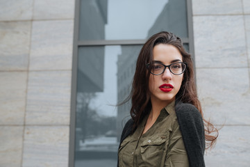 Fashion consept: beatiful young girl with long hair, glasses, red lips standing near modern wall wearing in green suit and grey jeans. She wanted talking on phone.