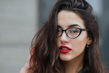 Fototapeta premium Fashion consept: beatiful young girl with long hair, glasses, red lips standing near modern wall wearing in green suit and grey jeans. She wanted talking on phone.