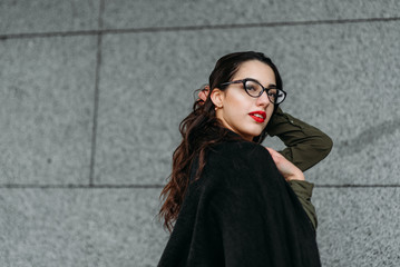 Naklejka premium Fashion consept: beatiful young girl with long hair, glasses, red lips standing near modern wall wearing in green suit and grey jeans. She wanted talking on phone.