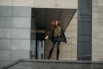 Fashion consept: beatiful young girl with long hair, glasses, red lips standing near modern wall wearing in green suit and grey jeans. She wanted talking on phone.