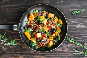 fried potatoes in a pan with tomatoes, mushrooms, olives, greens decorated with sprigs rosemary on a wooden table vegetarian food Top view