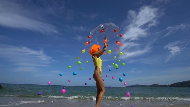 Sexy woman posing with colored balls attached to umbrella during summer day over sea and sky background. Creative look of woman in yellow swimsuit, orange wig and white sunglasses - slow motion video