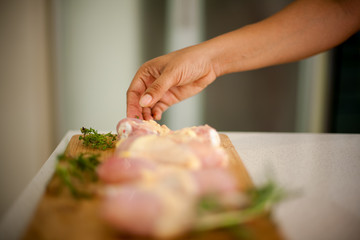 African American woman reaches for a piece of chicken, her hand in focus. In the foreground  blurred shot of drumsticks on a cutting board with rosemary and thyme leaves. .