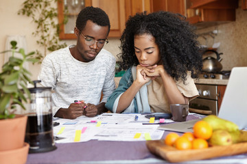 Young African couple doing paperwork together, sitting at kitchen table with lots of papers, calculator and laptop, looking frustrated. Dark-skinned family calculating domestic budget at home