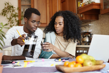 Young dark-skinned man in spectacle looking thoughtful holding pot, pouring coffee to his upset wife while managing family finances together in kitchen, using laptop computer and calculator