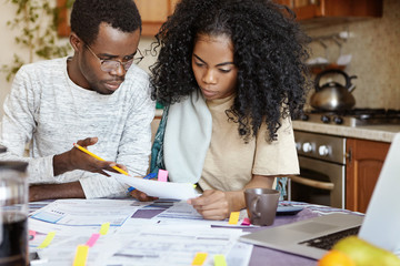 Indignant African husband gesturing with pencil, reproaching his wife for doing mistake while calculating bills, woman looking at piece of paper with serious frustrated expression. People and finances