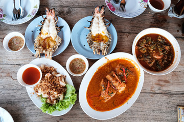 Many of Thai food on the wooden table include Fried squid with garlic, Grilled giant river prawn, Fried fish in spicy curry sauce and Spicy and soup curry with shrimp and vegetable omelet