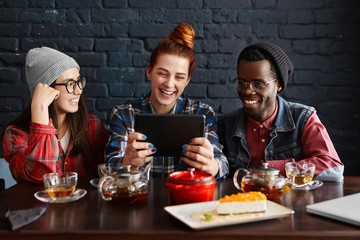 Multiethnic group of three young people enjoying online communication, using free wi-fi on generic touch pad, looking at screen with happy smiles while having lunch together at modern restaurant