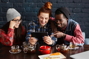 Teamwork, partnership and cooperation. Three ambitious young people brainstorming while discussing common project using touch pad at coffee shop. Multiethnic group working on electronic tablet