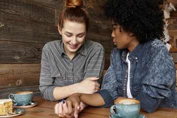 Beautiful Caucasian lesbian with hair bun relaxing at coffee shop with her stylish dark-skinned girlfriend in denim shirt. Happy homosexual couple enjoying great time together, holding hands