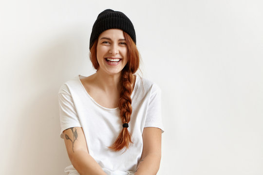 Indoor portrait of fashionable and cheerful young woman with braid wearing stylish black hat and white t-shirt smiling happily while posing in studio. Female fashion, beauty and advertisement concept