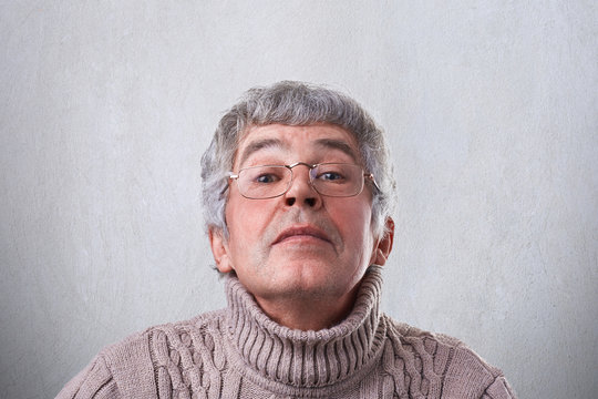 A close-up of funny grandfather in glasses looking happily through eyeglasses having fun. An elderly man having mysterious expression looking comically into camera while posing in studio