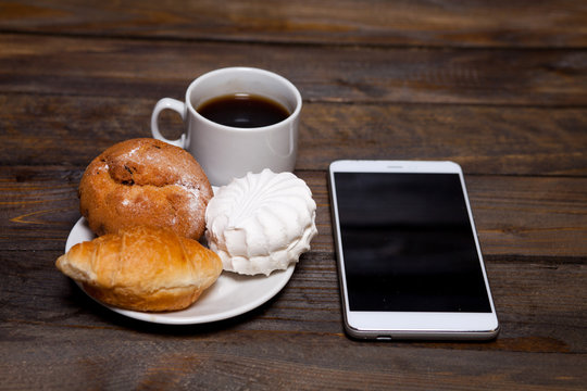 Coffee in white cup and cell phone with croissant, cake, marshmallows