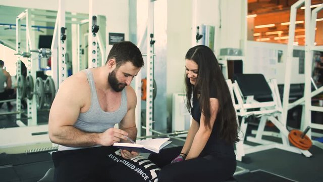 Trainer helps young strong brunette woman write training program in fitness club and gym center
