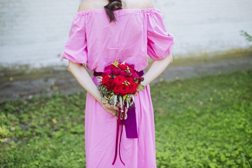Happy bride in a simple pink wedding dress holding a beautiful bouquet of red roses and green leaves behind her back. Woman in a stylish dress celebrating summer day wedding, blurred background