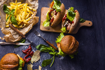 Fast food on the table: hot dog sausage and vegetables, burger with pork or beef and French fries with tomato sauce, basil leaves