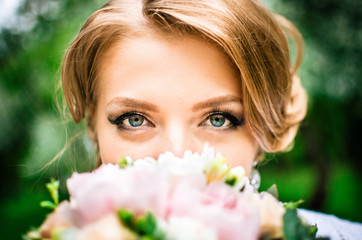 Fototapeta premium Bride with beautiful large green eyes and blonde hair looks into the camera hiding behind bouquet of flowers close up
