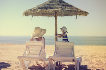Back view of joyful mother and daughter relaxing under beach umbrella sunny outdoors background