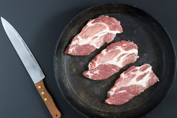 Fresh pork chops in cast iron skillet and knife on black background