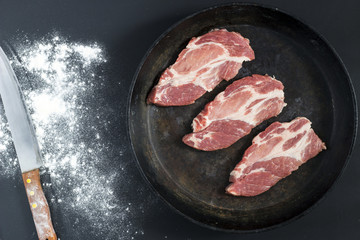 Fresh pork chops in cast iron skillet and knife on black background