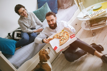Young hipster couple home eating pizza with laptop and dog