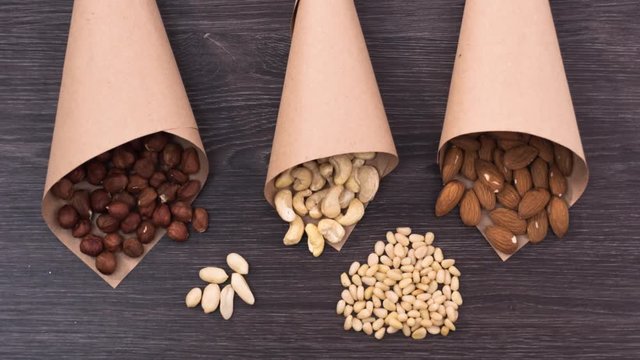 Three paper packages filled with nuts. Filbert, cashew and almonds fall in paper bags near a handful of pine nuts and peanuts. Stop motion
