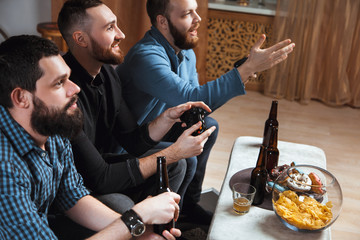 Men with a beard sitting on the couch at home with beer and chips with joysticks in hand playing computer video games. The concept of friendship, technology and weekend