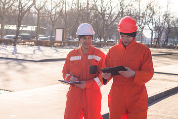 team of  young engineers discussing project and writing in the note book. The wear overalls and safety helmets. Street background