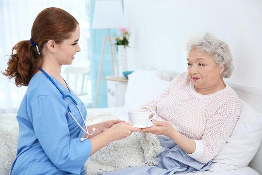 Nurse giving to elderly woman cup of tea in light room