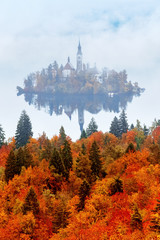 Pilgrimage Church of the Assumption of Maria at Blek lake in Slovenia. Autumn vertical scenery, forest with yellow foliage at foreground. Epic fall season landscape.