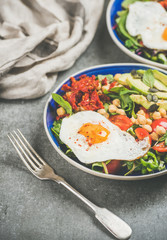 Healthy breakfast with fried egg, chickpea sprouts, seeds, fresh vegetables and greens in bowls over grey concrete background, selective focus. Clean eating, healthy lifestyle, vegetarian food concept