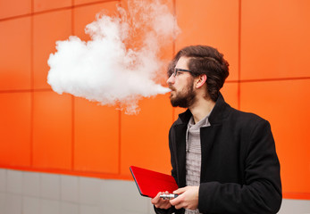 Young hipster man with a beard and glasses smokes an electronic cigarette. He holds a tablet pc....