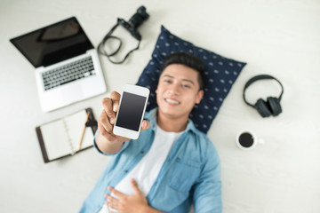 Top view of asian man lying on the floor with laptop, camera, tablet, coffee showing mobile phone screen