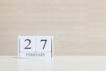 Closeup surface white wooden calendar with black 27 february word on blurred brown wood desk and wood wall textured background with copy space in selective focus at the calendar