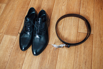 Men's classic black leather shoes and a leather belt with a buckle on wooden background.