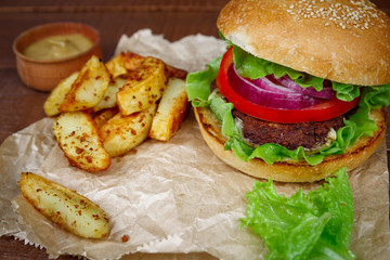 Tasty grilled beef burger with knife, lettuce and potatoes on dark wooden background. Big hamburger for lunch. Food photo.
