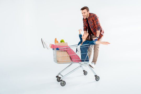 Excited Young Couple Riding On Shopping Cart With Grocery Bag On White