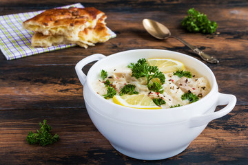 Avgolemono, chicken soup with egg-lemon sauce, rice and fresh parsley leaves in white bowl and freshly baked bread on wooden table.