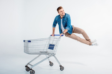 Excited young man jumping and having fun with shopping trolley on white
