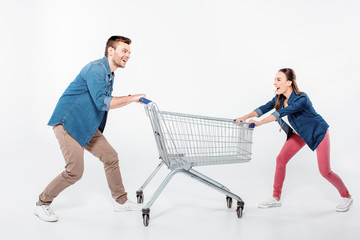 smiling couple pulling empty shopping cart and looking on each other on white