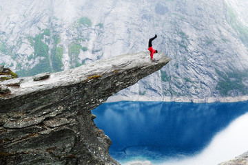 Young man climber in red jacket making intrepid handstand at the edge of famous Troltunga cliff over blue water of Ringedalsvatnet lake in canyon. Norway, summer scenery. Wanderlust concept.