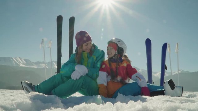 Mother and child girl are skiers sitting on snow against mountains. Laughing at camera