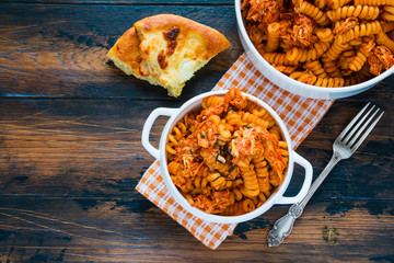 Fusilli pasta with chicken. Cooked in spicy sauce from tomatoes, onion, garlic, dried oregano and thyme, paprika and olive oil. Bread, white bowl and casserole on wooden brown rustic table. Top view.